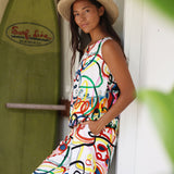A woman wearing a sleeveless white jumpsuit with a colorful abstract line pattern leans against a white door, with a green surfboard and a blurred green leaf in the background.
