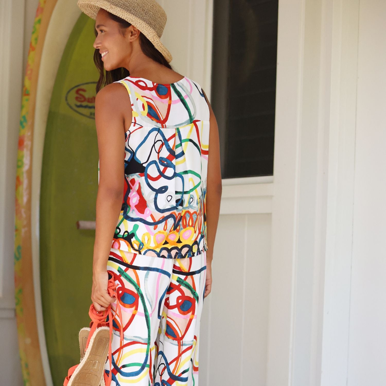 A woman wearing a sleeveless white jumpsuit with a colorful abstract graffiti-style print, a straw hat, and holding a straw hat, standing in front of a white door with a green surfboard in the background.
