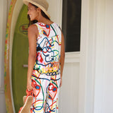 A woman wearing a sleeveless white jumpsuit with a colorful abstract graffiti-style print, a straw hat, and holding a straw hat, standing in front of a white door with a green surfboard in the background.