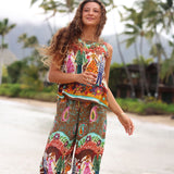 Une femme vêtue d'un haut sans manches à imprimé cachemire bohème coloré et d'un pantalon large assorti, se tient à l'extérieur sur un chemin de plage avec des palmiers et des montagnes floues en arrière-plan.