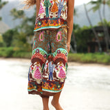Une femme vêtue d'un ensemble deux pièces imprimé et coloré, composé d'un haut sans manches et d'un pantalon à jambes larges, se tient à l'extérieur, près d'une plage ou de l'eau, avec un paysage tropical flou en arrière-plan.