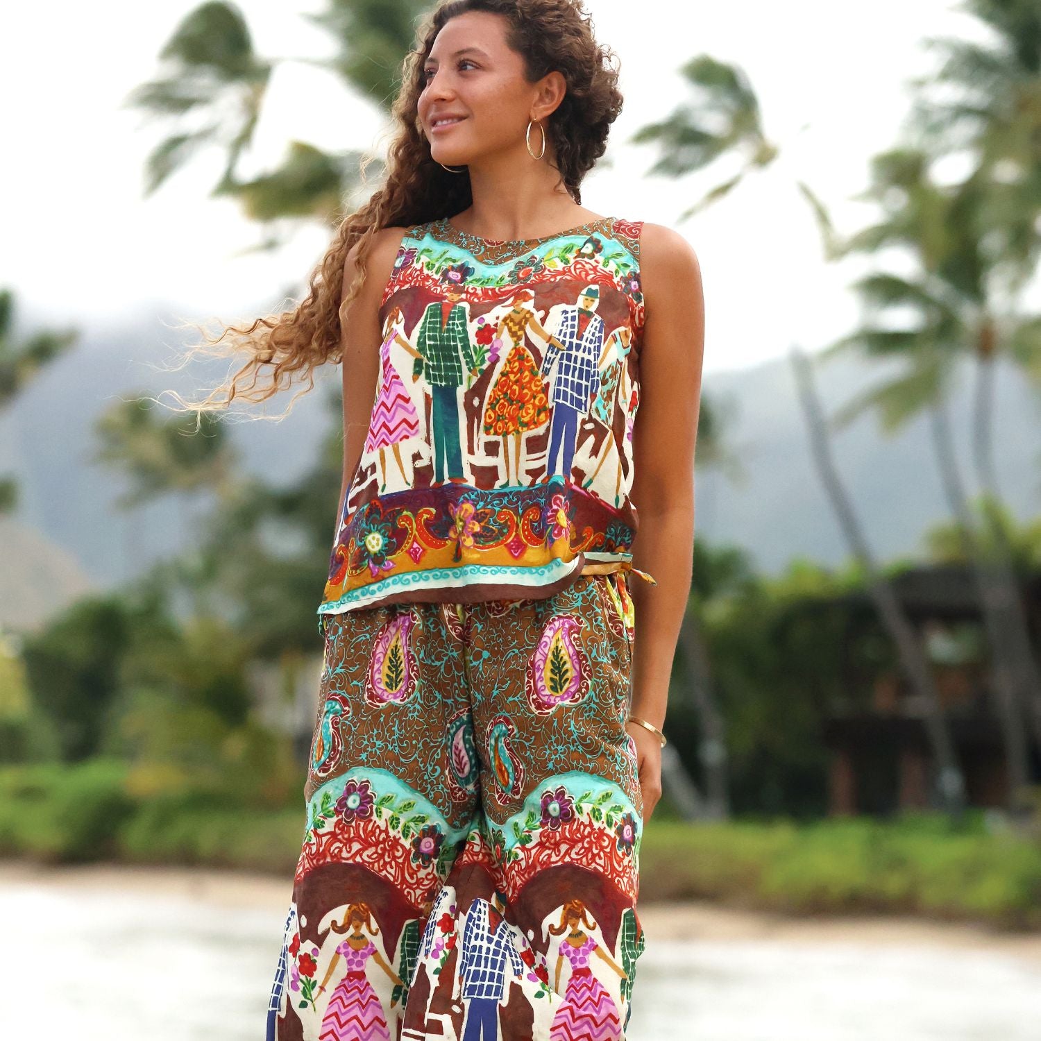 Une femme portant un débardeur sans manches et un pantalon large assorti, ornés d'un imprimé coloré et détaillé de style art populaire, se tient à l'extérieur devant un fond tropical flou avec des palmiers.