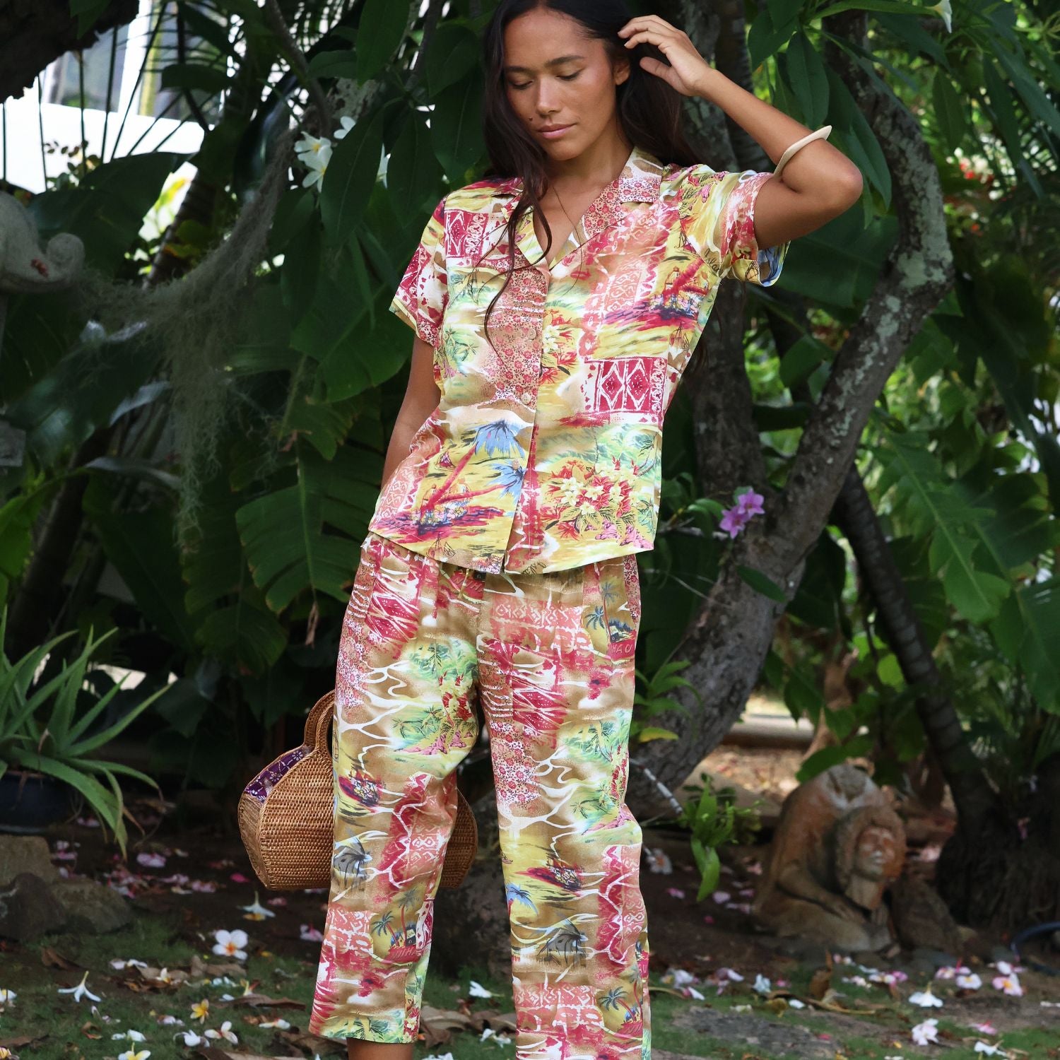 A woman wearing a matching short-sleeve button-up shirt and wide-leg pants pajama set with a colorful tropical patchwork print, standing in a lush, green outdoor garden setting.