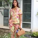 A woman stands outdoors in front of a white building with shuttered windows, wearing a matching short-sleeve button-up shirt and pants set in a bright multicolor floral print. She is holding a woven straw handbag with a colorful scarf tied to the handle.