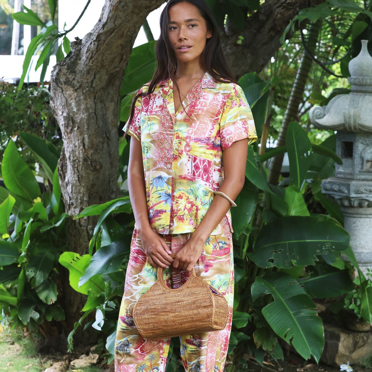 A woman stands outdoors in a tropical garden setting, wearing a matching two-piece pajama set with a vibrant multicolor abstract print. The set includes a short-sleeve button-up shirt and matching pants.