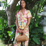 A woman stands outdoors in a tropical garden setting, wearing a matching two-piece pajama set with a vibrant multicolor abstract print. The set includes a short-sleeve button-up shirt and matching pants.