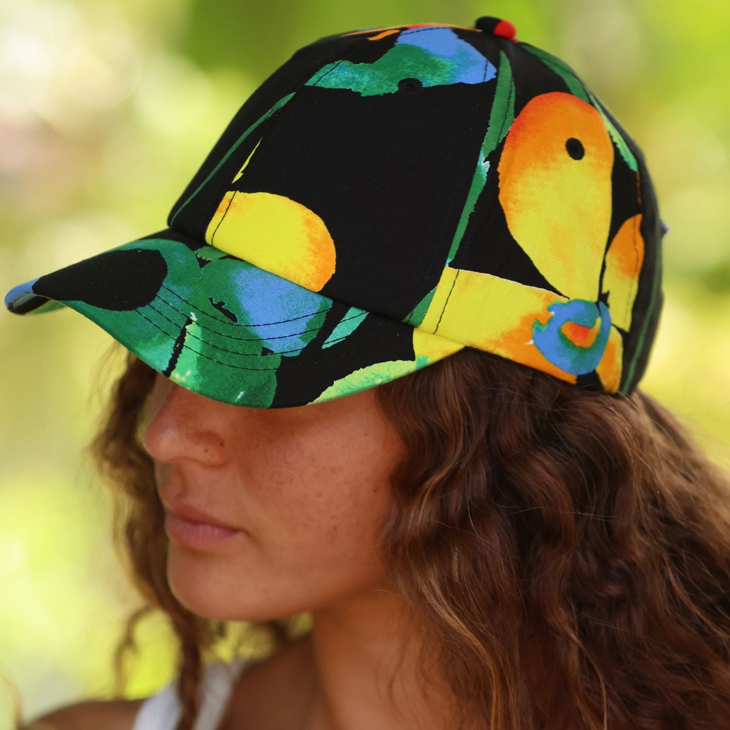 A close-up side profile of a woman wearing a black baseball cap with a vibrant, multicolored tropical leaf and bird pattern in green, yellow, orange, and blue, set against a soft, blurred green background.