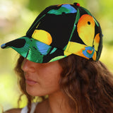 A close-up side profile of a woman wearing a black baseball cap with a vibrant, multicolored tropical leaf and bird pattern in green, yellow, orange, and blue, set against a soft, blurred green background.