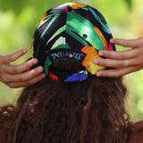 A person viewed from behind wearing a colorful baseball cap with hands adjusting the back strap. The cap has a black base with a vibrant abstract pattern in green, orange, yellow, blue, and pink, and a black adjustable strap with 'SAMSWORLD' embroidered in multicolored letters.