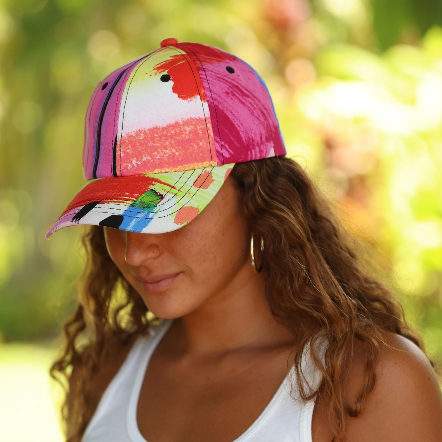 A woman wearing a vibrant, multicolored abstract-print baseball cap in an outdoor setting with a soft green bokeh background.