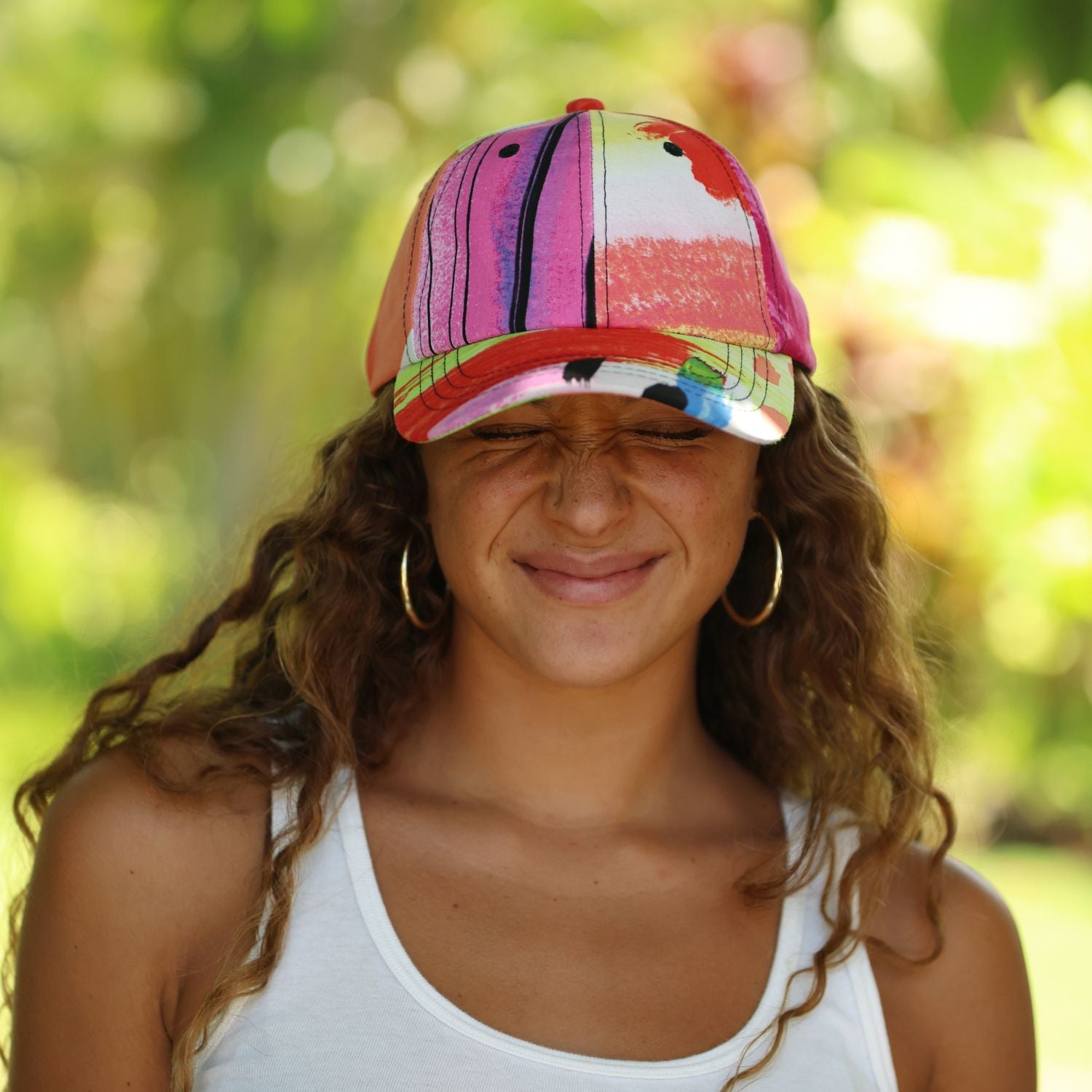 A woman outdoors wearing a colorful baseball cap with vertical stripes and abstract paint-like patterns in pink, orange, green, black, and white, along with gold hoop earrings and a white tank top.