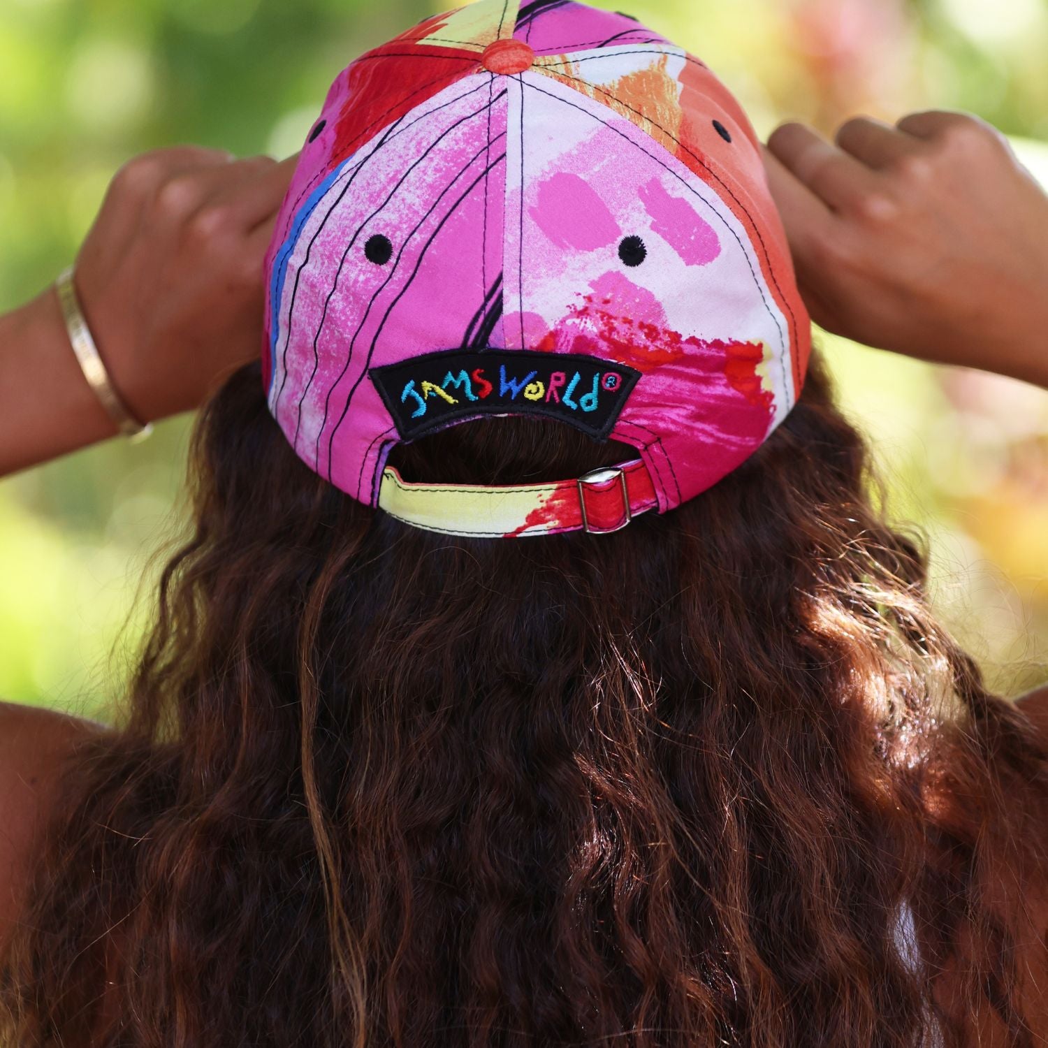 A person viewed from behind wearing a colorful baseball cap with hands adjusting it. The cap has a vibrant pink, orange, and white abstract paint-splatter pattern and a black brand patch reading 'SAM'S WORLD' on the back.