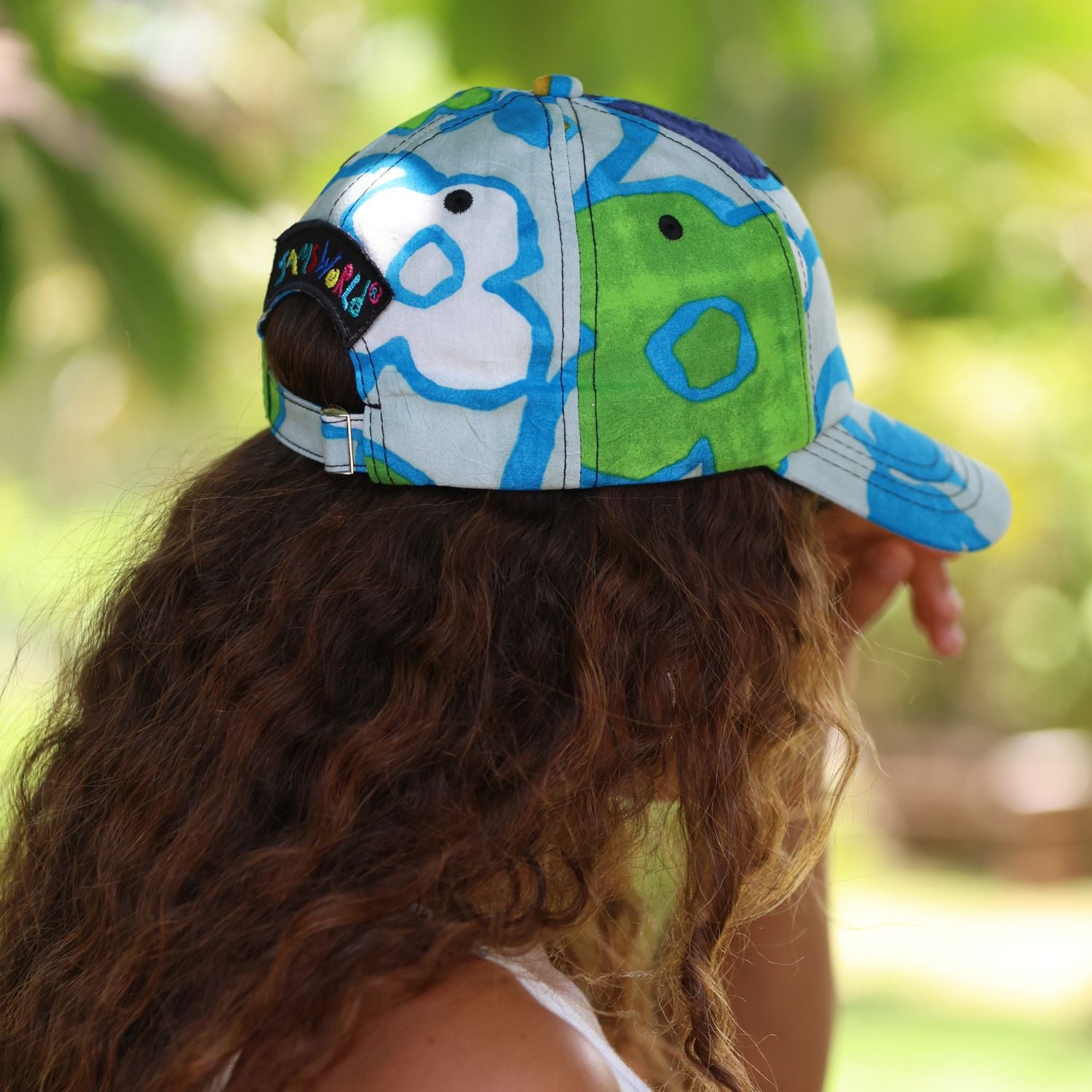 A person outdoors wearing a baseball cap with a blue, green, and white abstract pattern. The back of the cap has an adjustable black strap with colorful embroidered letters, and the background is a blurred green outdoor setting.
