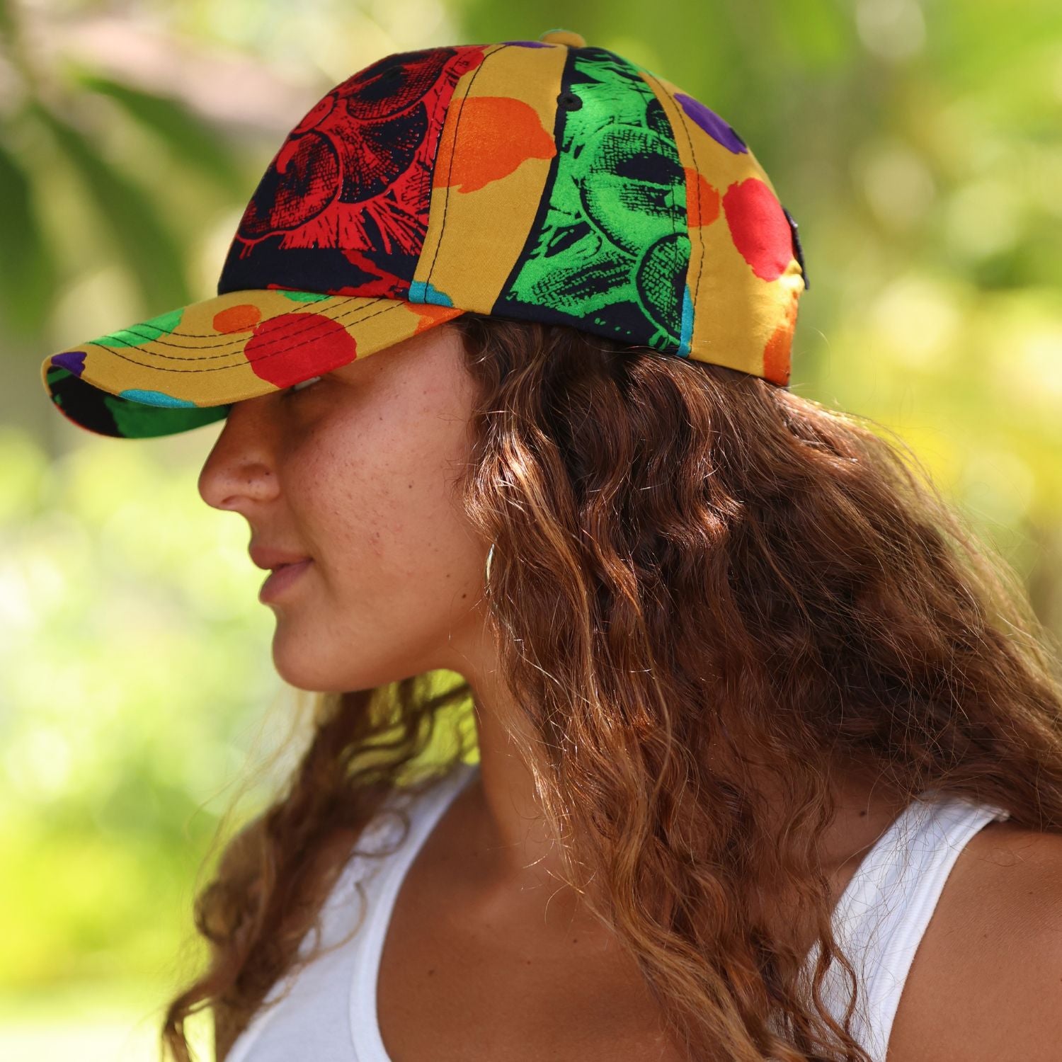 A woman in profile wearing a colorful patterned baseball cap with yellow, green, red, and black panels, photographed outdoors against a blurred green background.