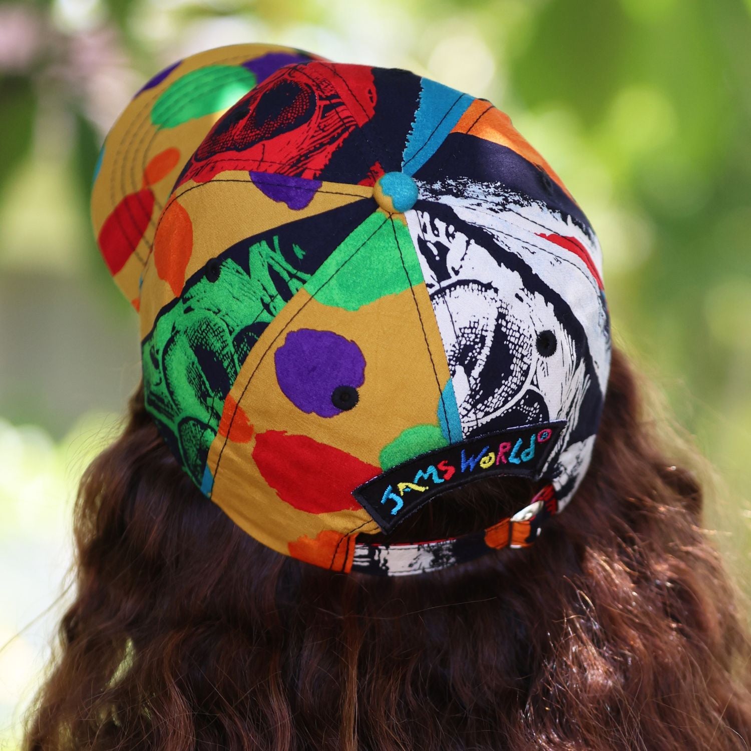 A colorful patterned baseball cap worn by a person, shown from the back. The cap features a vibrant, multicolored abstract print with yellow, green, red, blue, black, and white sections, and a black patch on the back with the text 'SMShWORLD'.