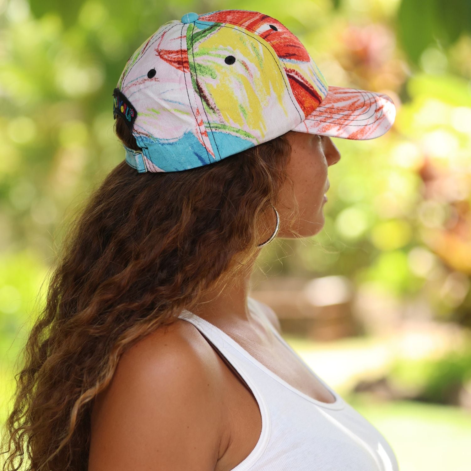 A woman in profile wearing a white baseball cap with a colorful abstract print in yellow, red, and blue, set against a blurred green outdoor background.
