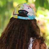 A person viewed from behind wearing a colorful baseball cap with a visible 'Sandy World' patch on the back, set against a blurred green outdoor background.