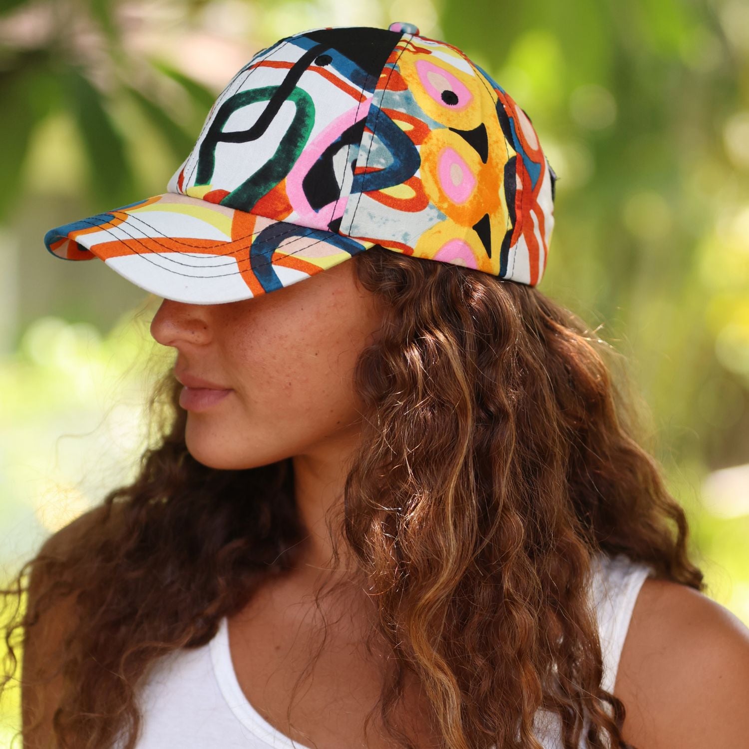 A woman in profile wearing a baseball cap with a vibrant, multicolored abstract pattern. The background is a soft-focus green outdoor setting.