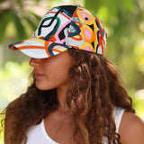 A woman in profile wearing a baseball cap with a vibrant, multicolored abstract pattern. The background is a soft-focus green outdoor setting.