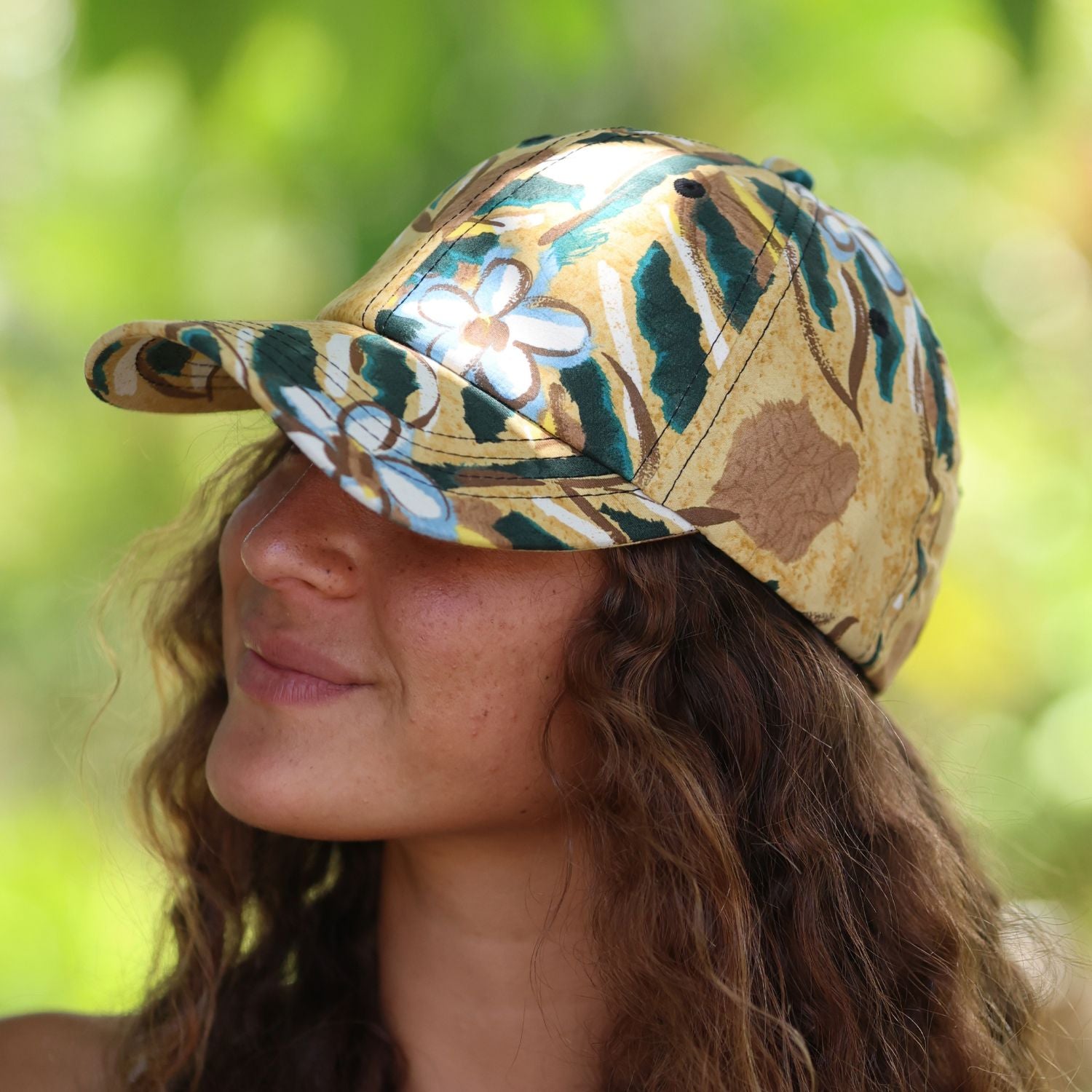 A woman wearing a baseball cap with a tropical floral pattern in beige, green, brown, and white, photographed outdoors against a blurred green background.