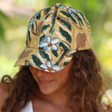 A woman wearing a baseball cap with a hand-painted style floral pattern in green, brown, and light blue on a tan background, photographed outdoors with a soft green bokeh background.