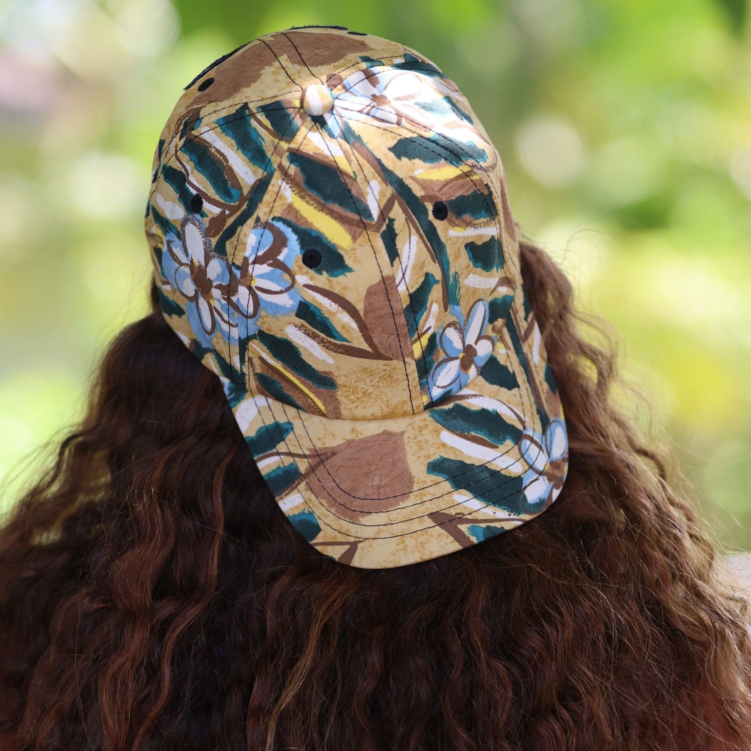 A person viewed from behind wearing a baseball cap with a tropical floral and leaf pattern in beige, brown, green, and white, set against a blurred green outdoor background.