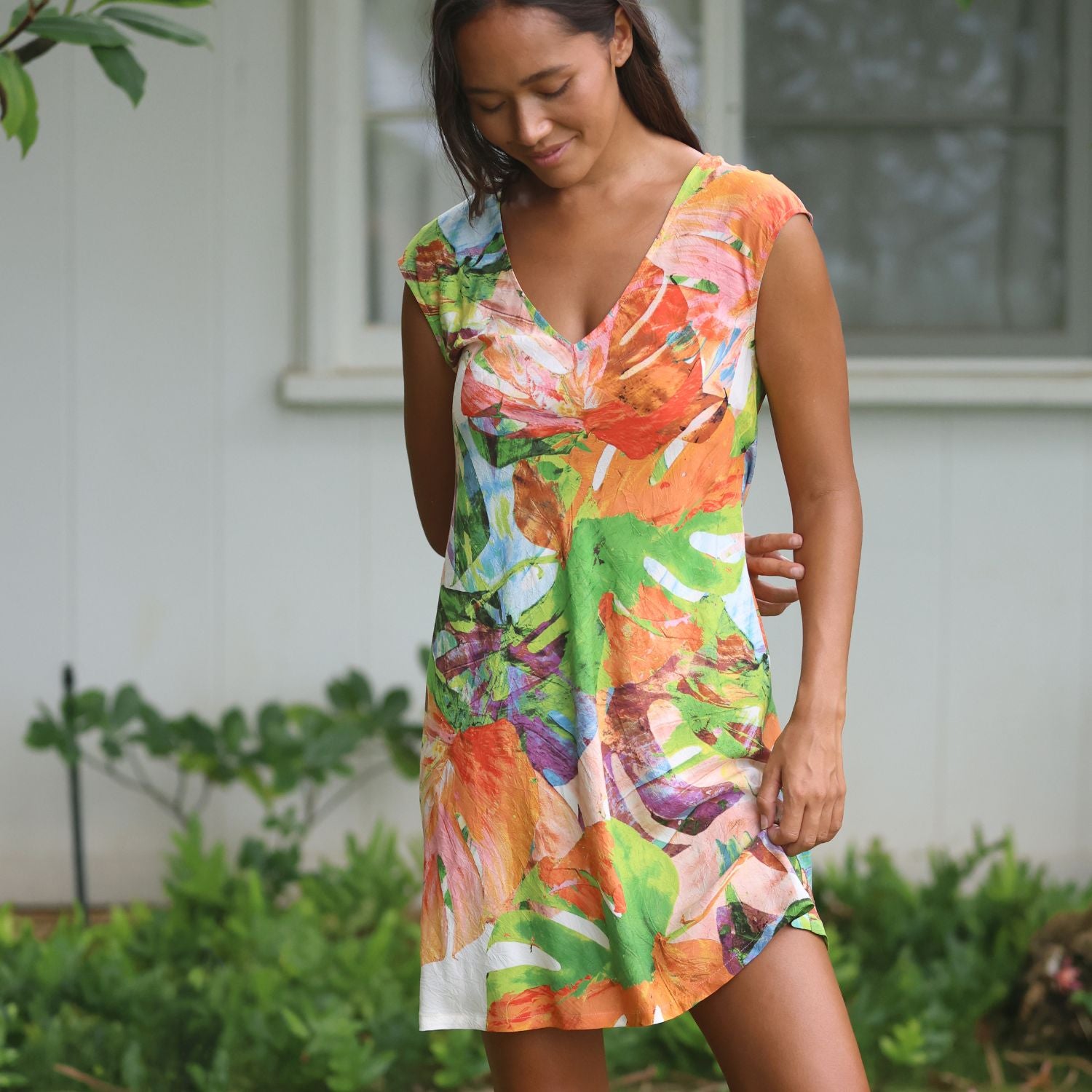 Una mujer con un vestido sin mangas con escote en V y un colorido estampado floral abstracto en naranja, verde, rosa y blanco, de pie al aire libre frente a la pared blanca de una casa y plantas verdes.