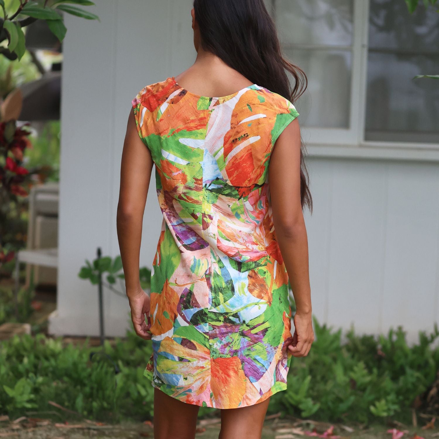Una mujer vista de espaldas con un vestido sin mangas hasta la rodilla con un vibrante estampado floral abstracto multicolor en naranja, verde, blanco y morado, sobre un fondo exterior de una casa blanca y vegetación.