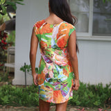 A woman viewed from behind wearing a sleeveless, knee-length dress with a vibrant multicolor abstract floral print in orange, green, white, and purple, set against an outdoor background of a white house and greenery.