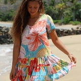 A woman wearing a colorful tropical floral tiered dress, standing on a sandy beach with water and greenery in the background.