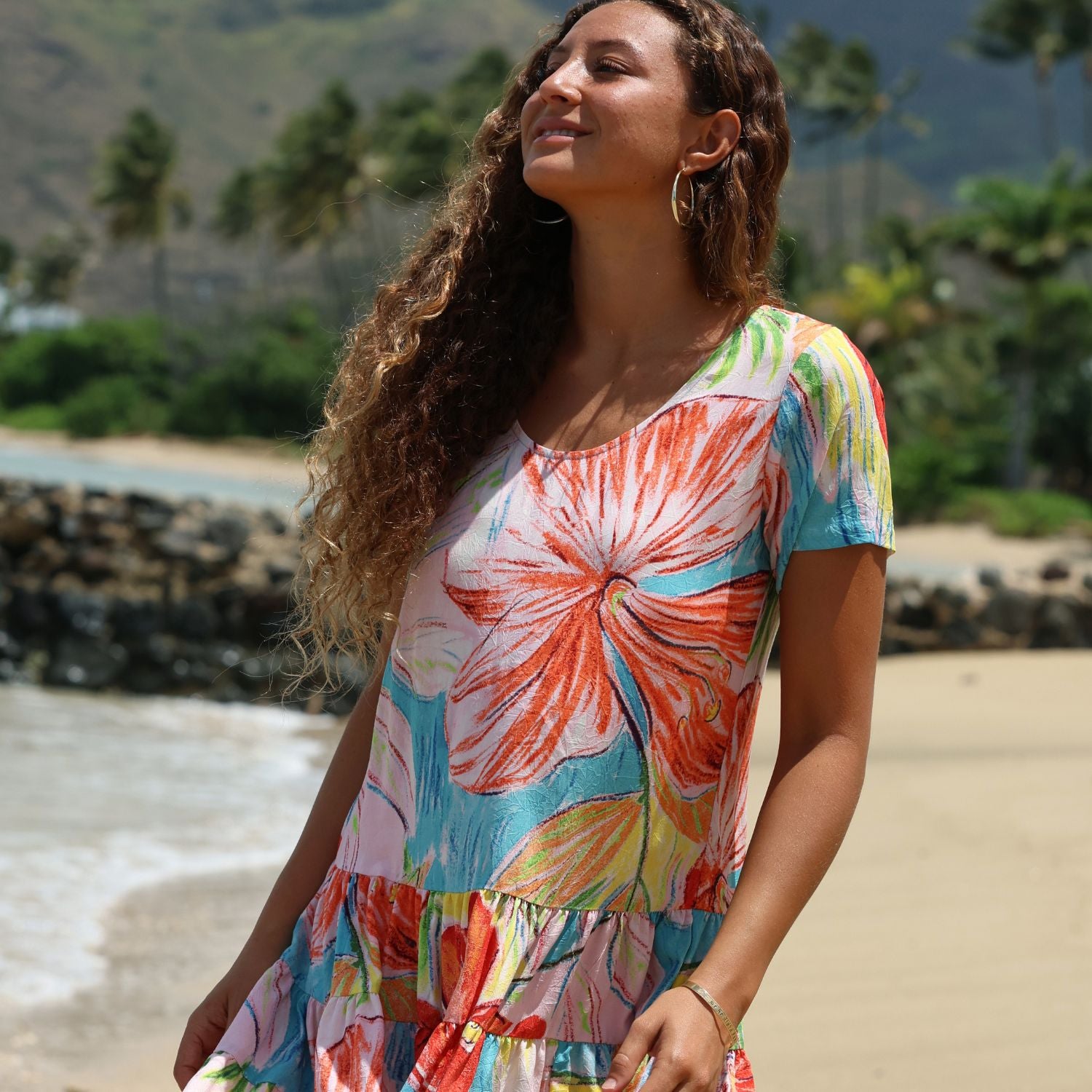 A woman with long curly brown hair stands on a sandy beach wearing a short-sleeve tiered dress with a colorful floral print. The dress features a large orange and red flower on a white background with light blue, green, and yellow accents.