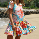 A woman wearing a tiered V-neck dress with a colorful tropical floral print, standing on a sandy beach with water and greenery in the background.
