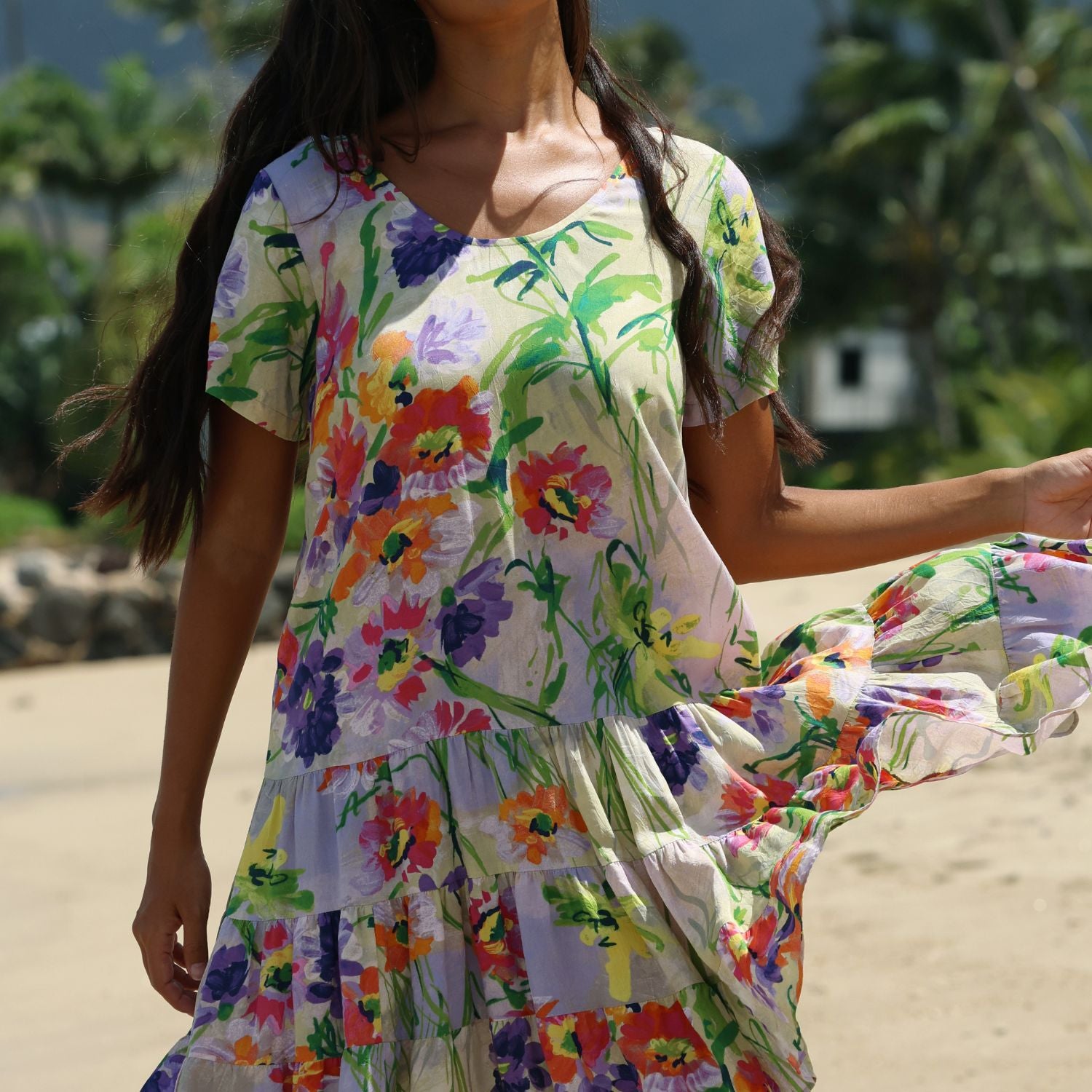 A woman wearing a short-sleeve tiered dress with a vibrant multicolor floral print, standing outdoors on a sandy beach with palm trees in the background.