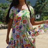 A woman wearing a short-sleeve tiered dress with a vibrant multicolor floral print, standing outdoors on a sandy beach with palm trees in the background.