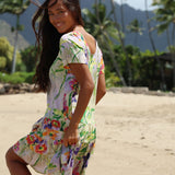 A woman wearing a short-sleeve, scoop-neck dress with a colorful tropical floral print, standing on a sandy beach with palm trees and mountains in the background.