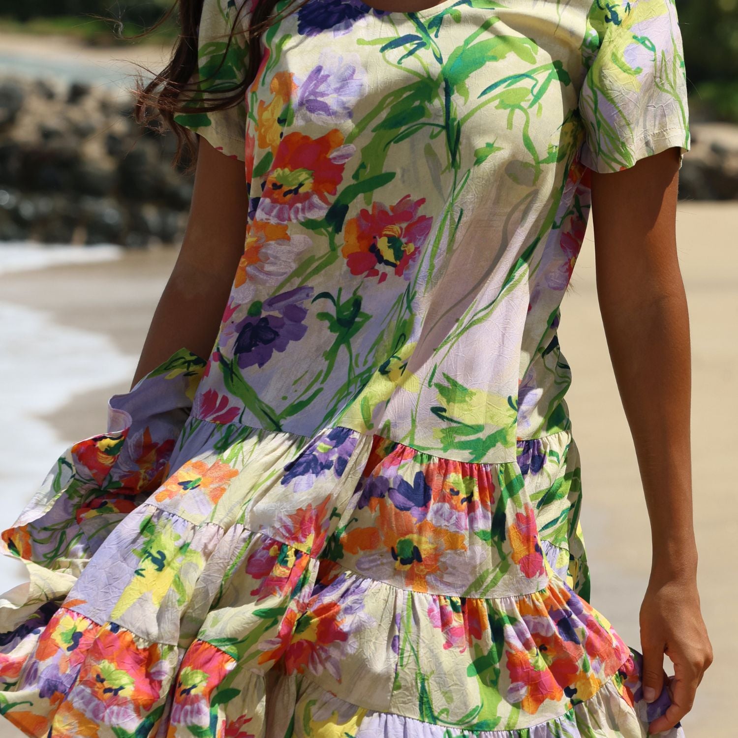 A woman wearing a tiered short-sleeve dress with a colorful floral print, standing on a sandy beach with water and rocks blurred in the background.