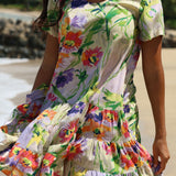A woman wearing a tiered short-sleeve dress with a colorful floral print, standing on a sandy beach with water and rocks blurred in the background.