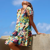 A woman wearing a colorful, short-sleeved tiered dress with a vibrant abstract floral print, leaning against a concrete wall under a bright blue sky.
