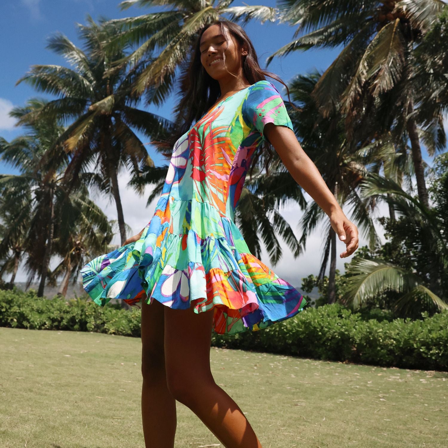 A woman wearing a colorful tropical print short-sleeve dress stands outdoors on a grassy lawn with palm trees and blue sky in the background.