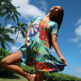 A woman wearing a colorful tropical print tiered dress poses outdoors against a background of blue sky and palm trees.