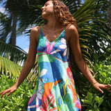 A woman wearing a colorful tropical floral print sleeveless dress stands outdoors among lush green palm leaves and foliage, with her arms outstretched and her head tilted back in a sunny natural setting.