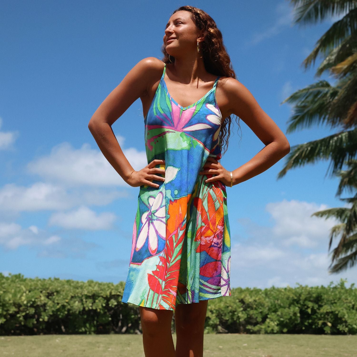 A woman wearing a colorful tropical floral print sleeveless dress stands outdoors with her hands on her hips against a blue sky and palm tree background.