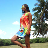 A woman wearing a colorful tie-dye sleeveless mini dress and yellow sunglasses, posing outdoors on a grassy lawn with palm trees and a blue sky in the background.