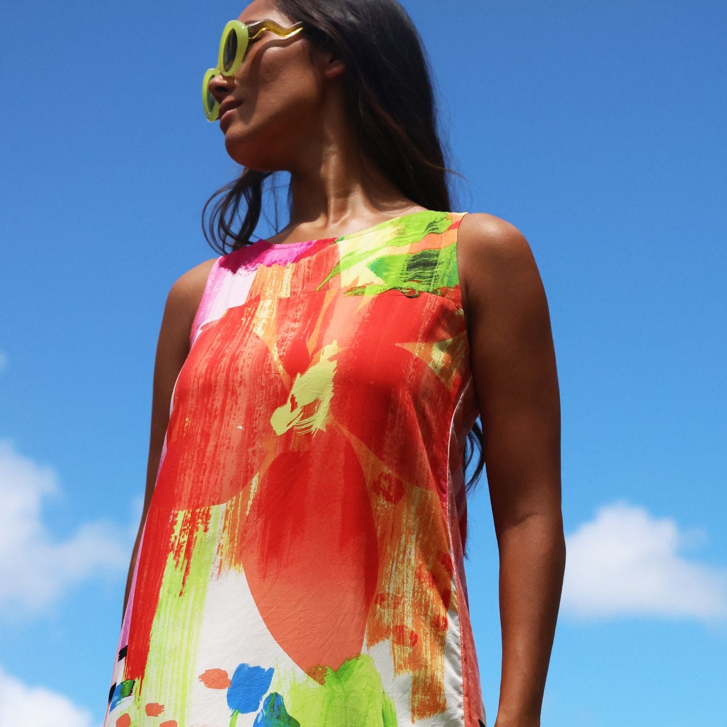 A woman wearing a sleeveless, colorful abstract-print dress and yellow sunglasses, photographed outdoors against a bright blue sky.