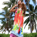 A woman wearing a vibrant, multicolored abstract-print sleeveless dress stands outdoors in front of palm trees, looking upwards with her hands near her head.