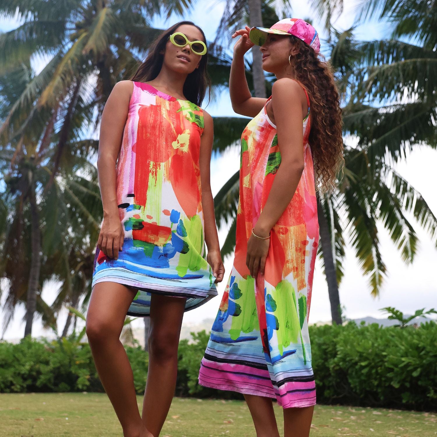 Two women wearing matching sleeveless tropical abstract print dresses in an outdoor park setting with palm trees. The dresses feature a vibrant mix of pink, orange, green, blue, and white paint strokes with striped borders.