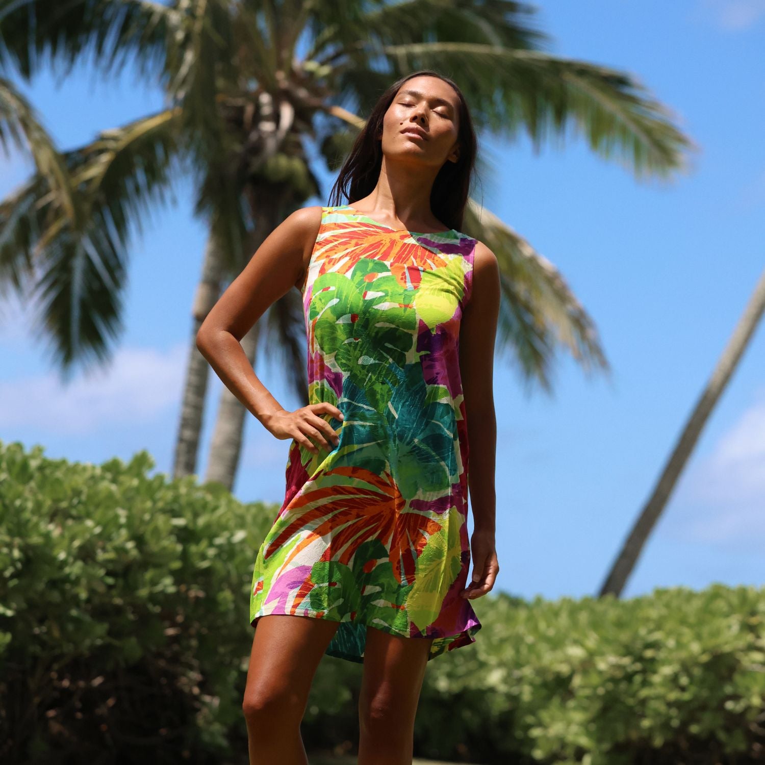 A woman wearing a sleeveless, knee-length dress with a vibrant tropical floral print in green, yellow, orange, and purple, posing outdoors against a background of palm trees and blue sky.