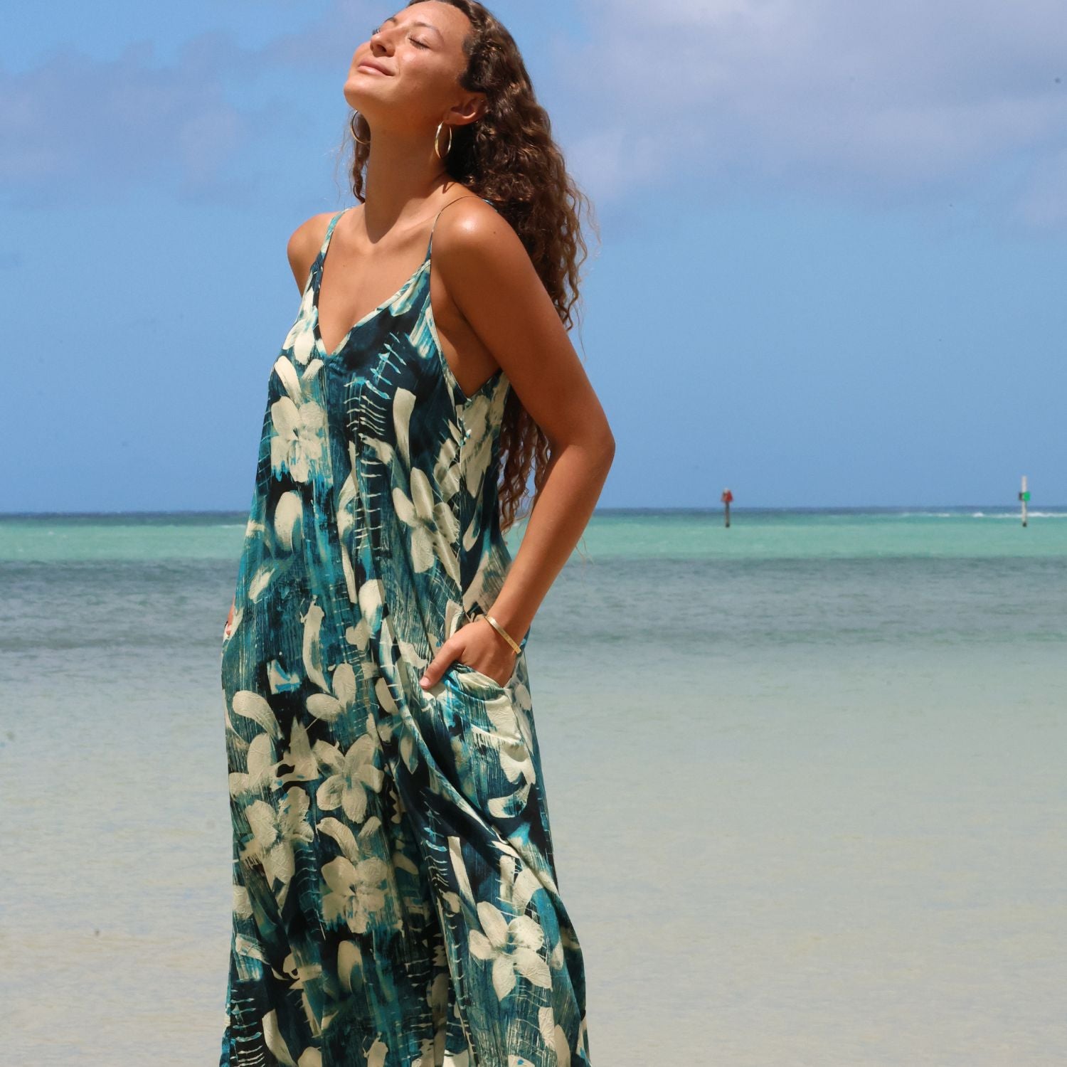 A woman wearing a teal and white floral maxi dress stands on a sandy beach with the ocean and blue sky in the background.