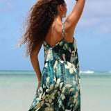 A woman on a beach wearing a sleeveless backless dress with a teal, black, and cream tropical floral print. She is shown from behind with her hair blowing in the wind, with the ocean and blue sky in the background.