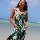 A woman wearing a sleeveless V-neck maxi dress with a teal, green, and cream abstract floral print, standing on a sunny beach with the ocean and blue sky in the background.